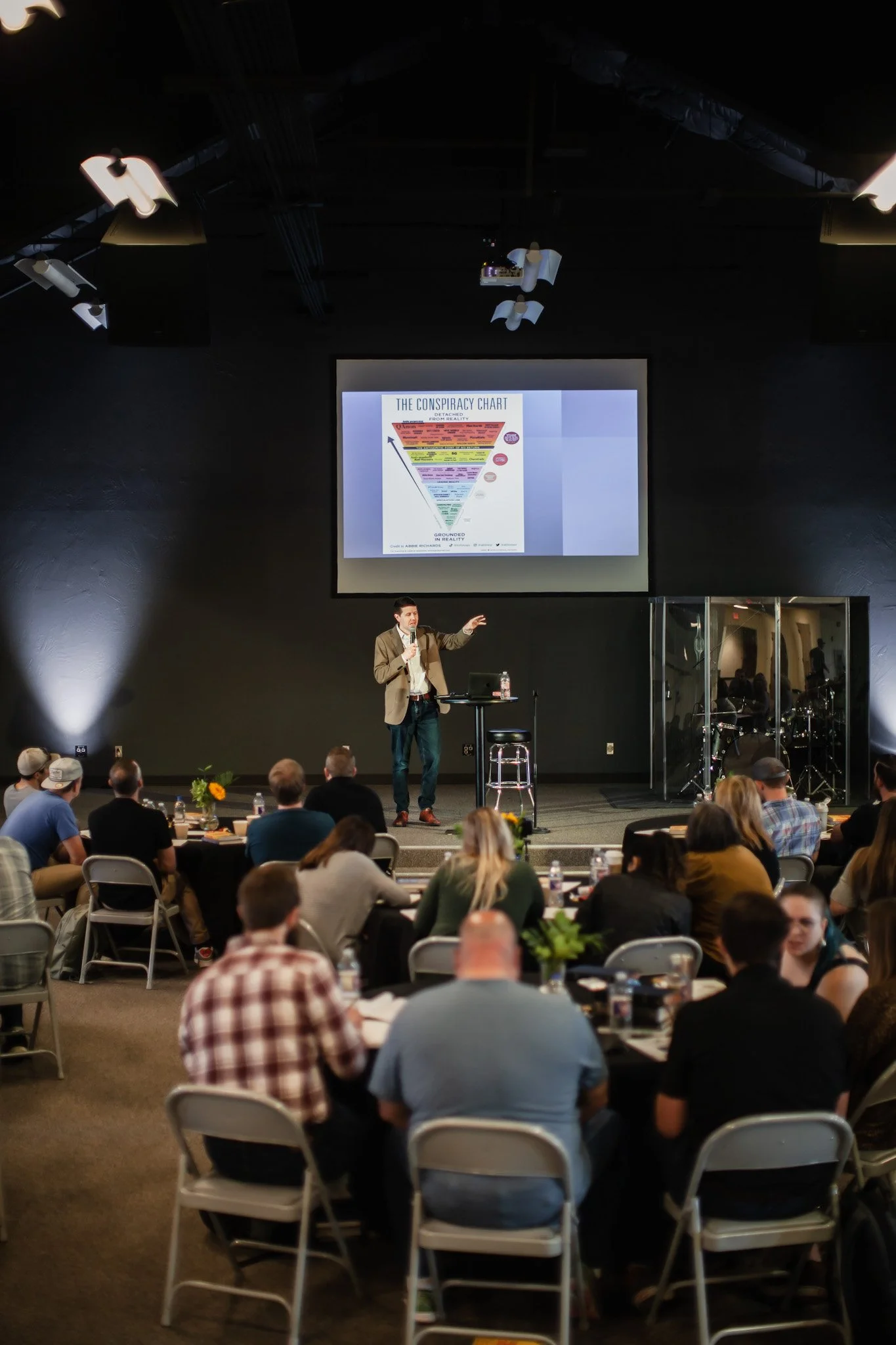 A man giving a presentation to an audience in a theater or conference room. The screen behind him displays a chart titled 'The Conspiracy Chart' with a funnel diagram. The audience is seated at round tables with water bottles and flower arrangements.