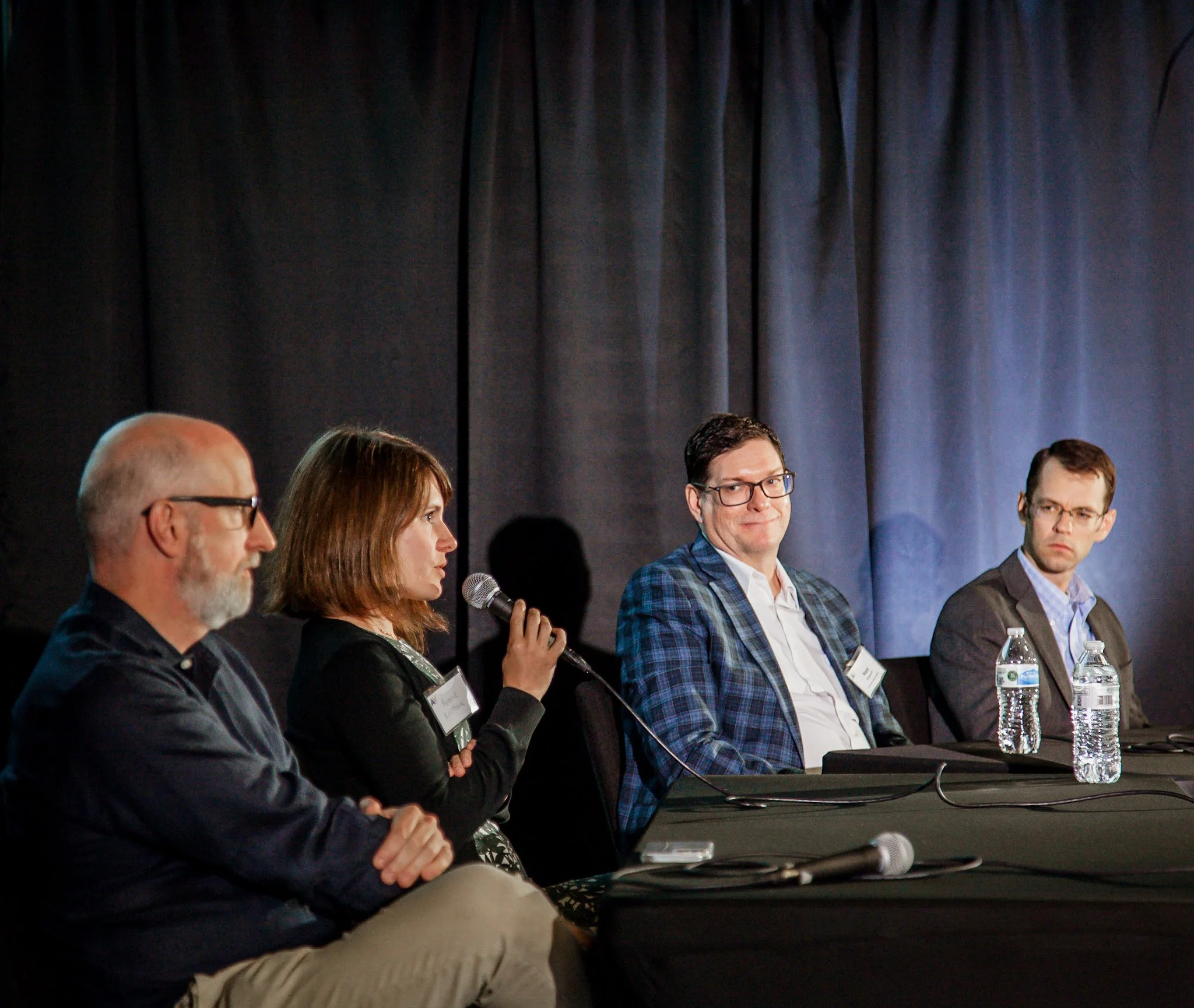 Four people seated at a panel discussion with a dark curtain backdrop, two of them holding microphones, and water bottles on the table.